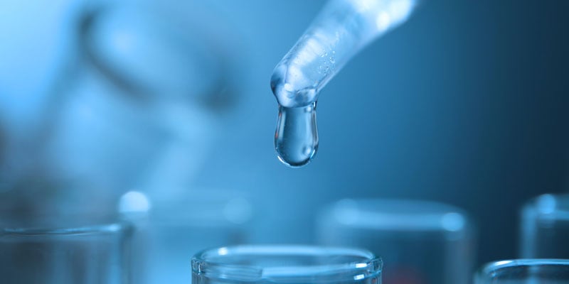 A glass dropper releases a single droplet above a clear vial in a blue-lit laboratory.