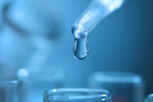 A glass dropper releases a single droplet above a clear vial in a blue-lit laboratory.